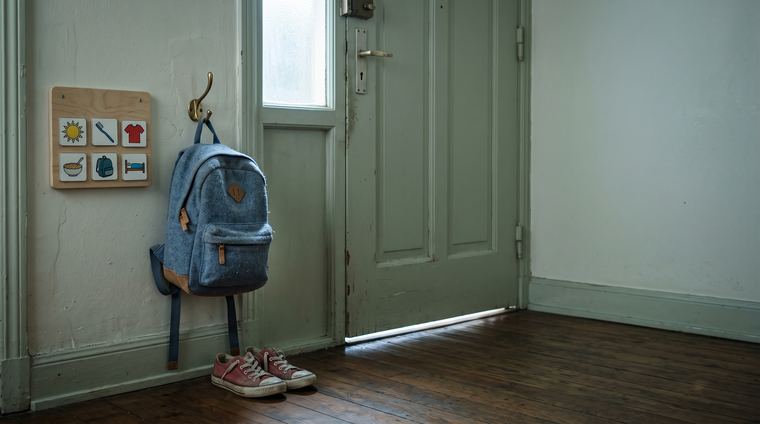 Cartable d'enfant accroché à un patère près de la porte d'entrée avec un planning visuel de pictogrammes au mur et les baskets posées : la rentrée d'un enfant TDAH ou TSA se prépare avant le jour J