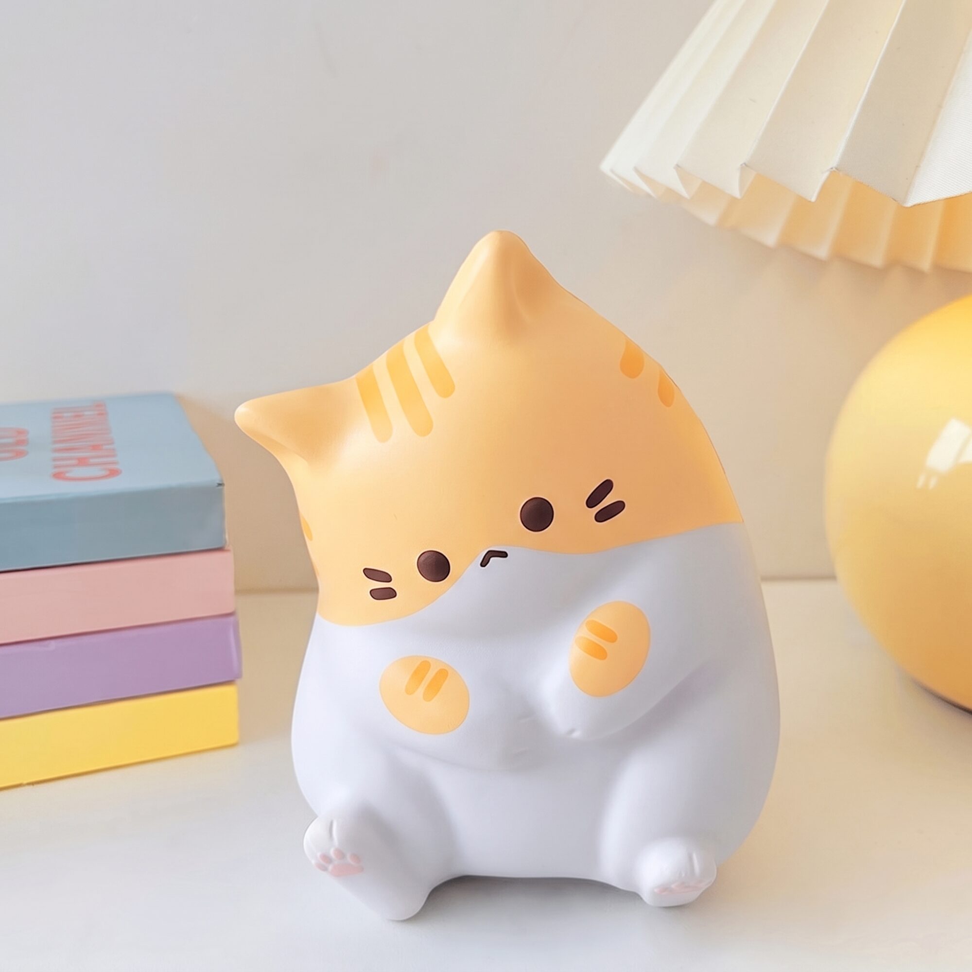 Veilleuse en forme de chat blanc et orange assise, à côté d'une pile de livres aux couleurs pastel