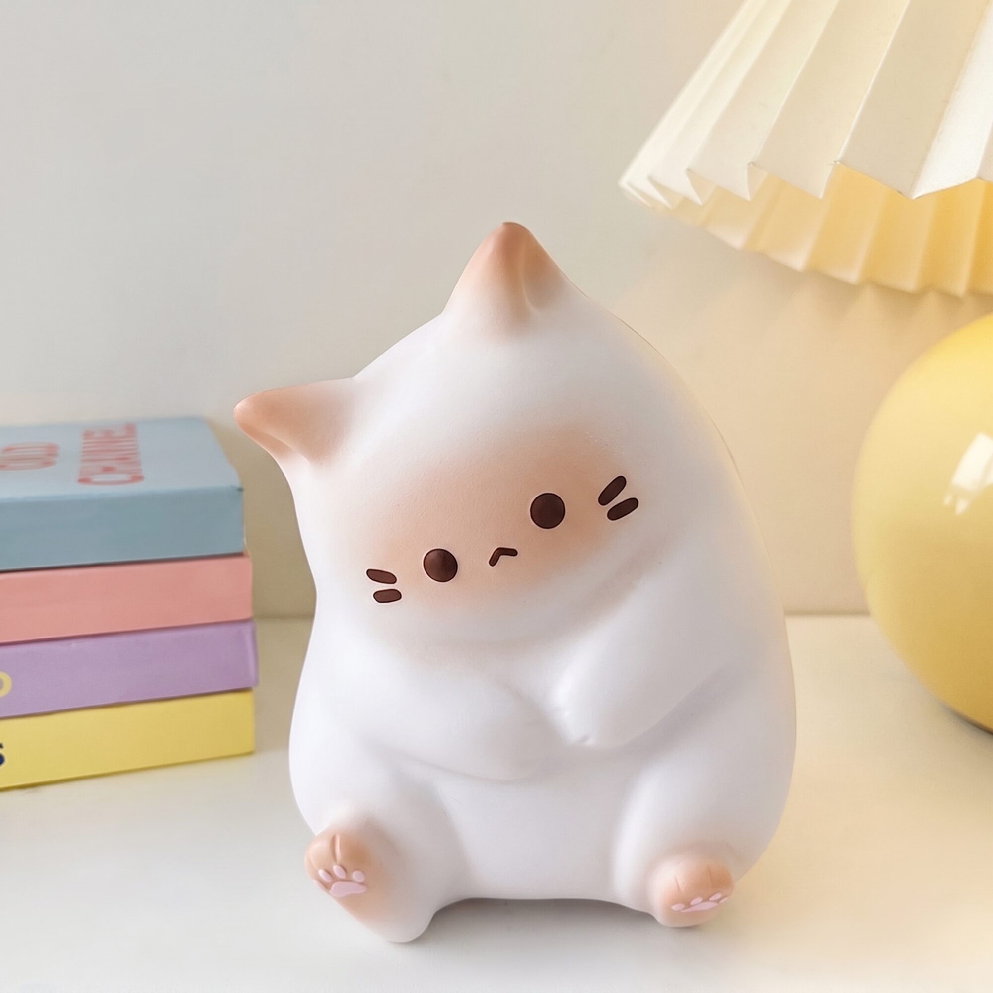 Veilleuse en forme de chat blanc aux oreilles et coussinets pêche, assise devant une pile de livres aux couleurs pastel