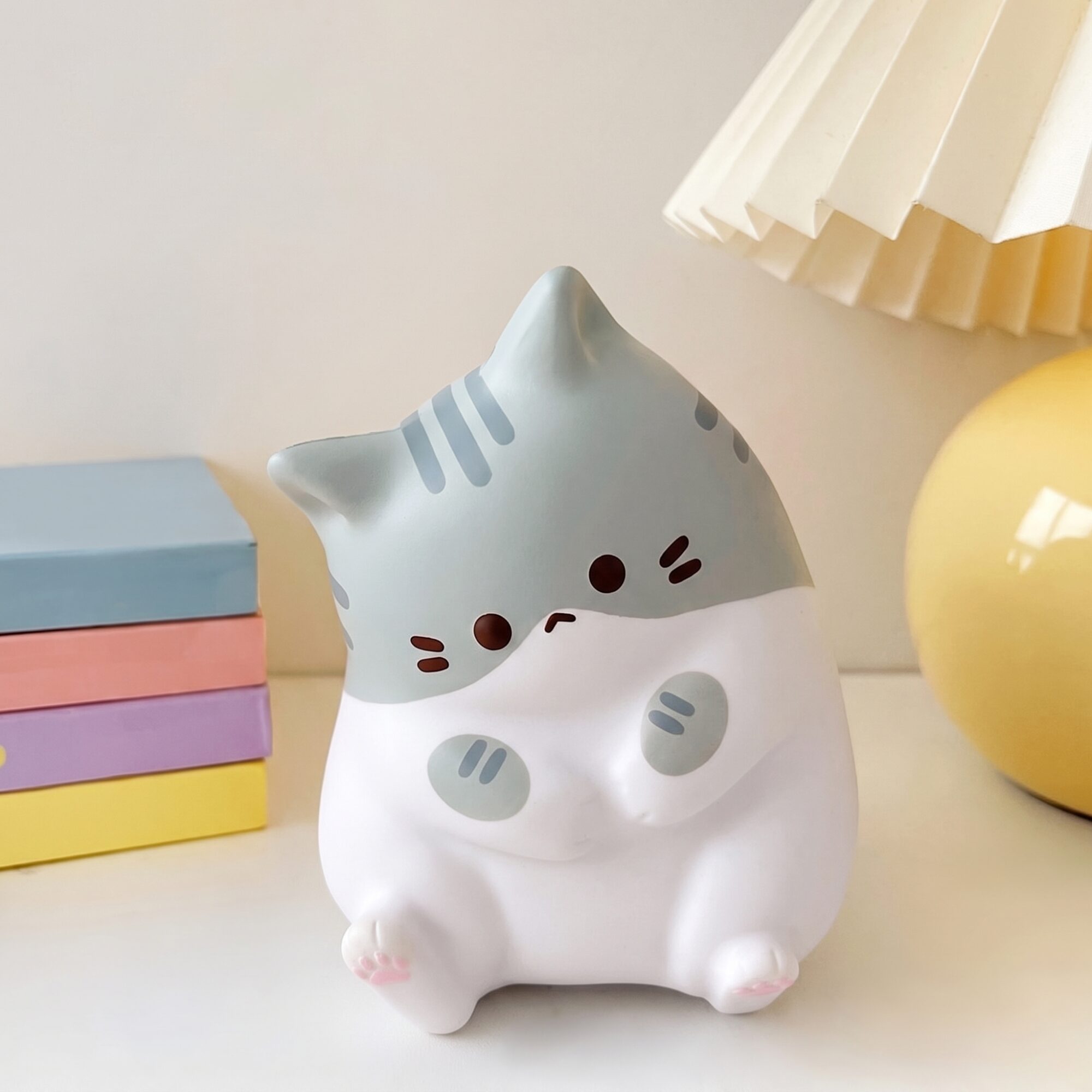 Veilleuse en forme de chat à tête grise rayée et corps blanc, assise devant une pile de livres aux couleurs pastel