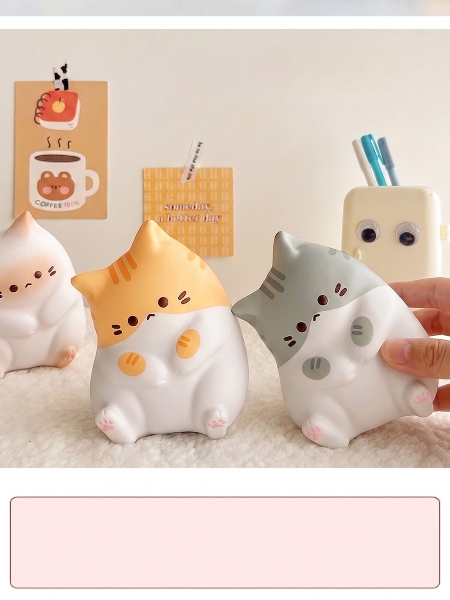 Trois veilleuses en forme de chats assis, blanche, orange et grise sur un tapis blanc, une main pressant celle à tête grise