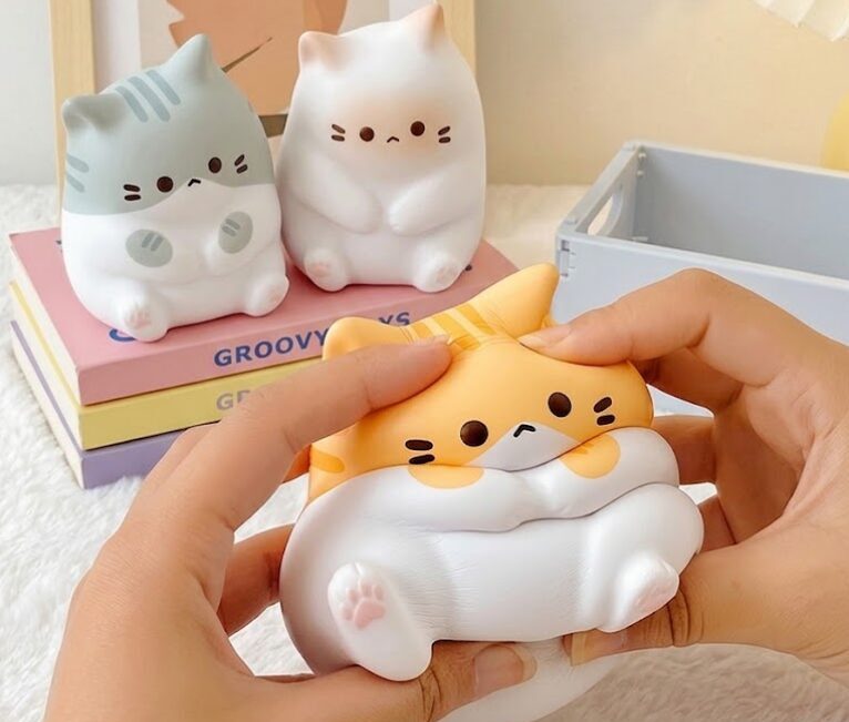 Mains pressant une figurine souple de chat blanc et orange qui se déforme, deux autres figurines de chats posées en arrière-plan sur des livres